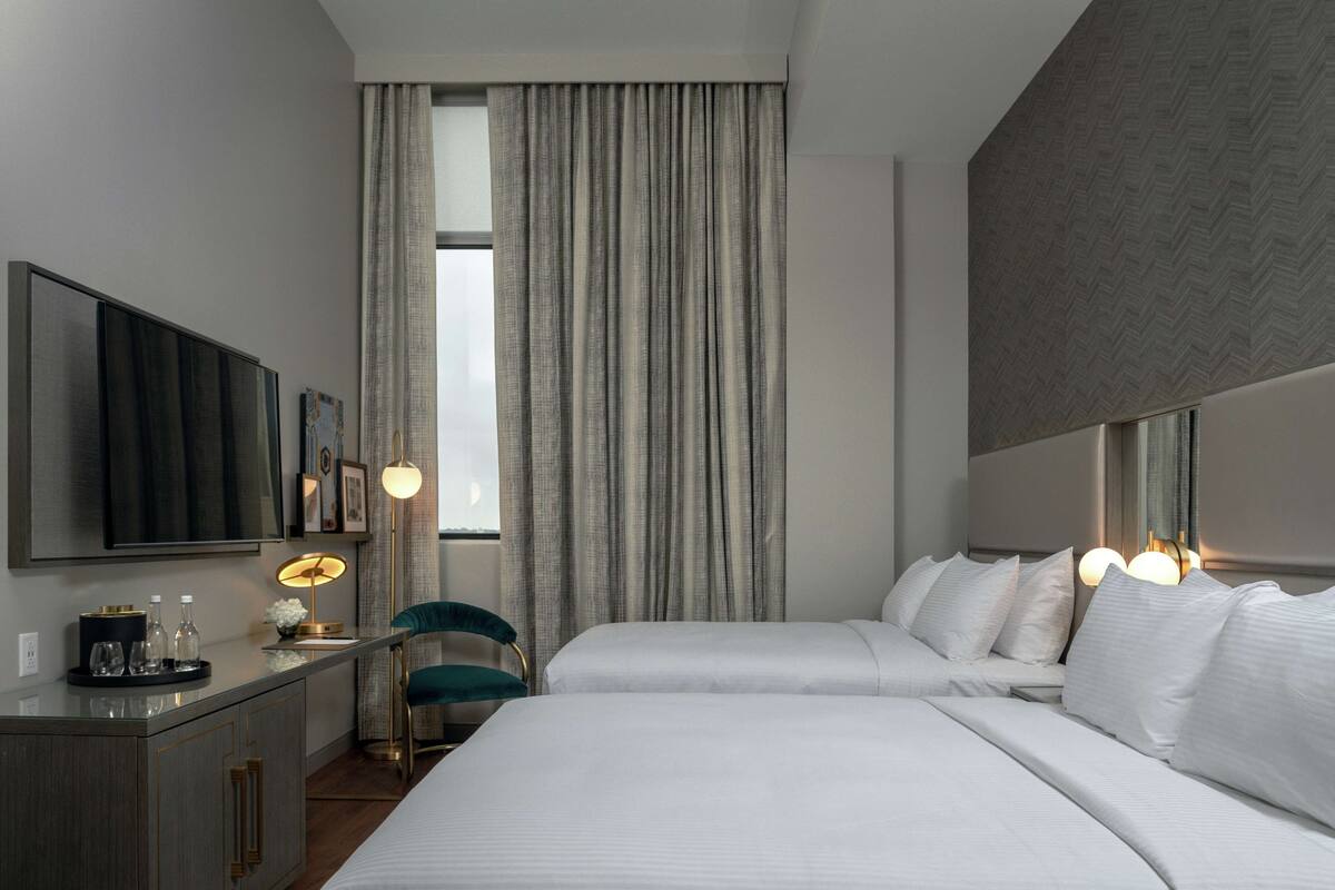 A modern hotel room with a large bed, a desk with a flat-screen TV, a chair, and a large window with curtains.