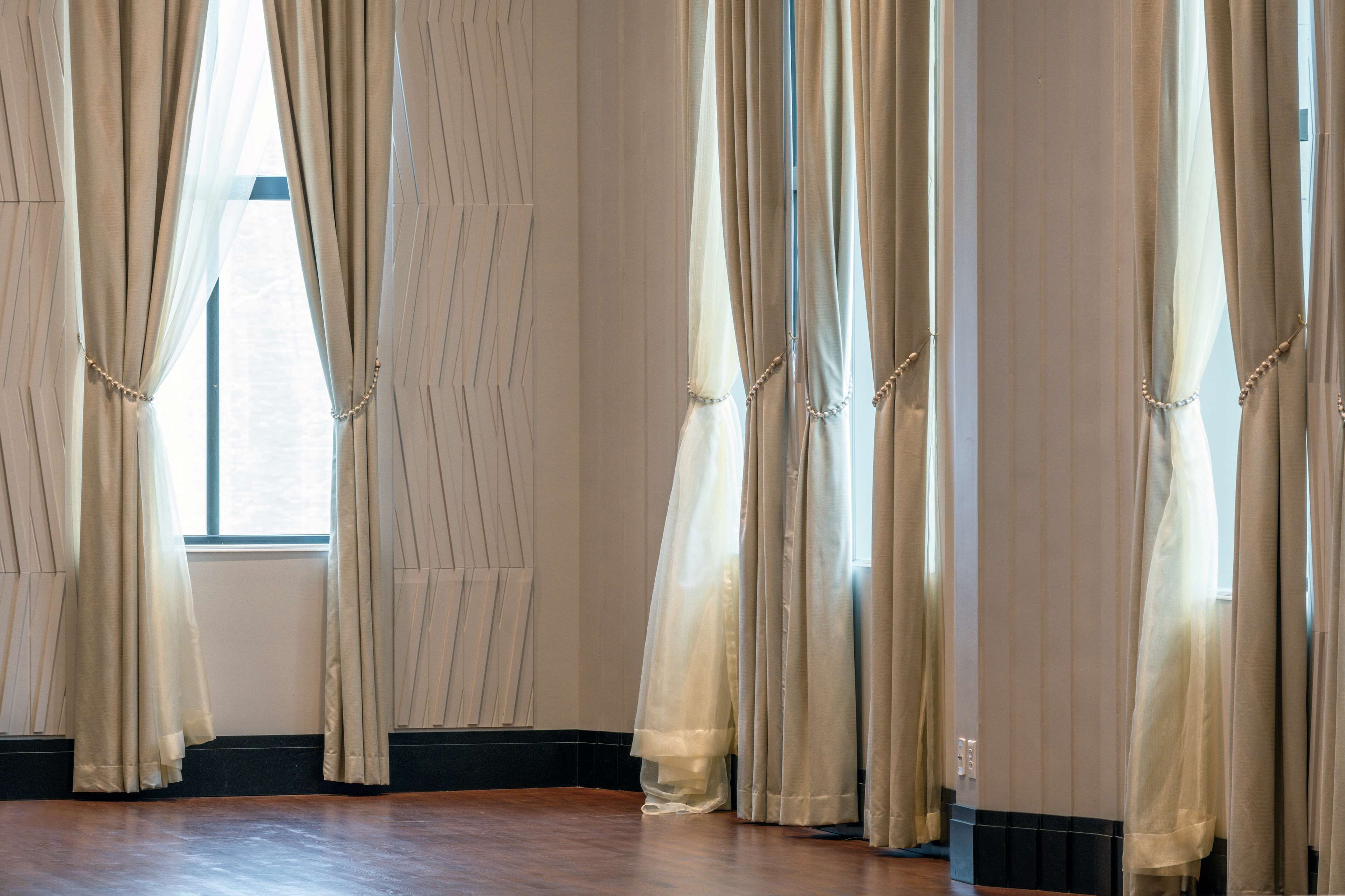 Una habitación con ventanas amplias, cortinas color beige y suelo de madera.