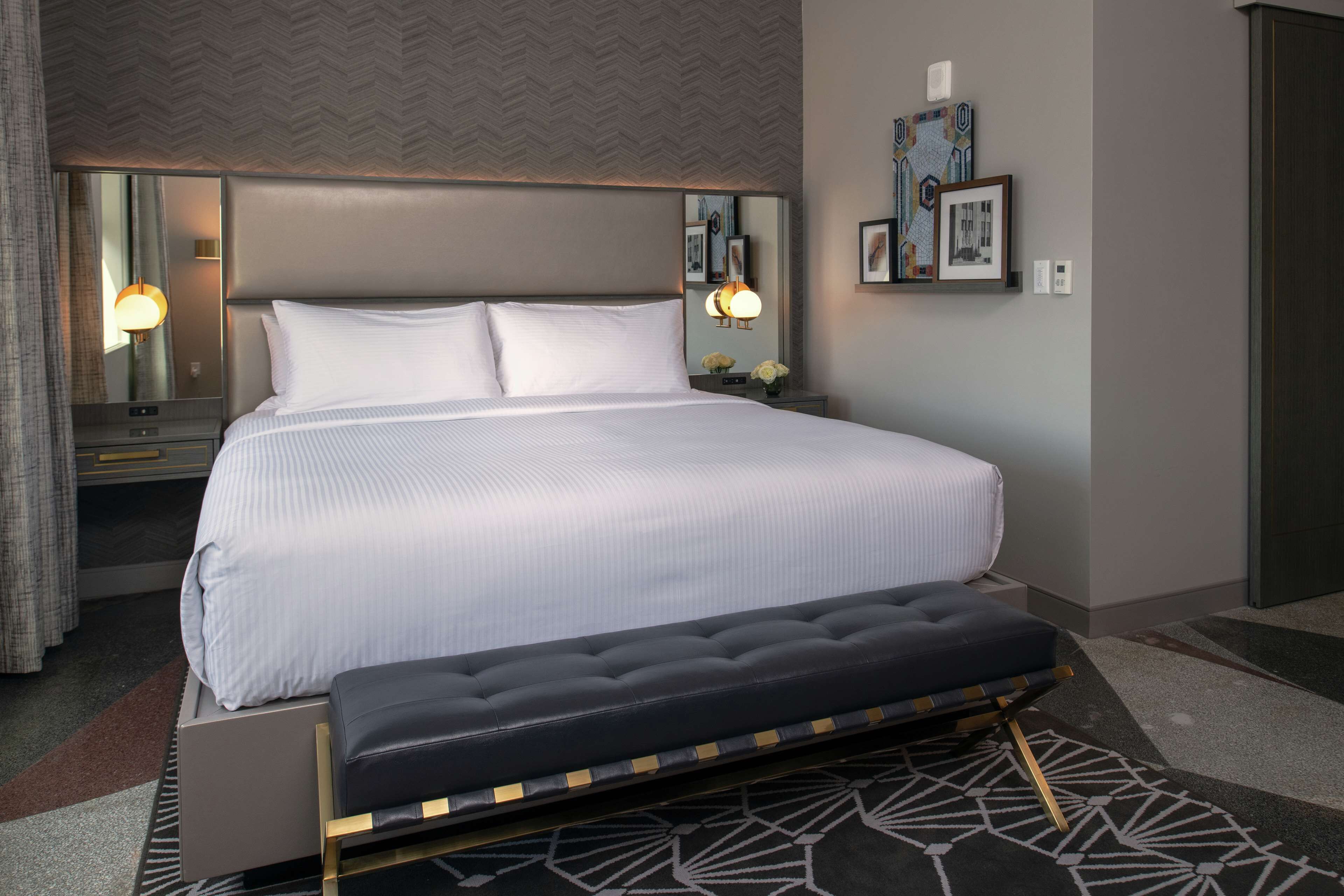 Una habitación de hotel moderna con una cama grande, una mesita de noche con lámpara, un banco y una alfombra estampada.