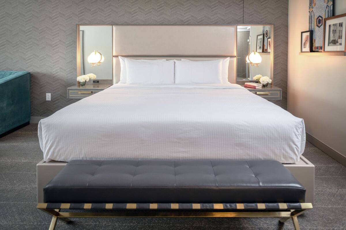 A neatly arranged hotel room with a large bed, bedside tables with lamps, a bench, and a herringbone-patterned wall.