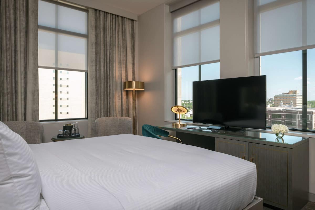 A hotel room with a large bed, a desk with a TV, a lamp, and a view of the city.