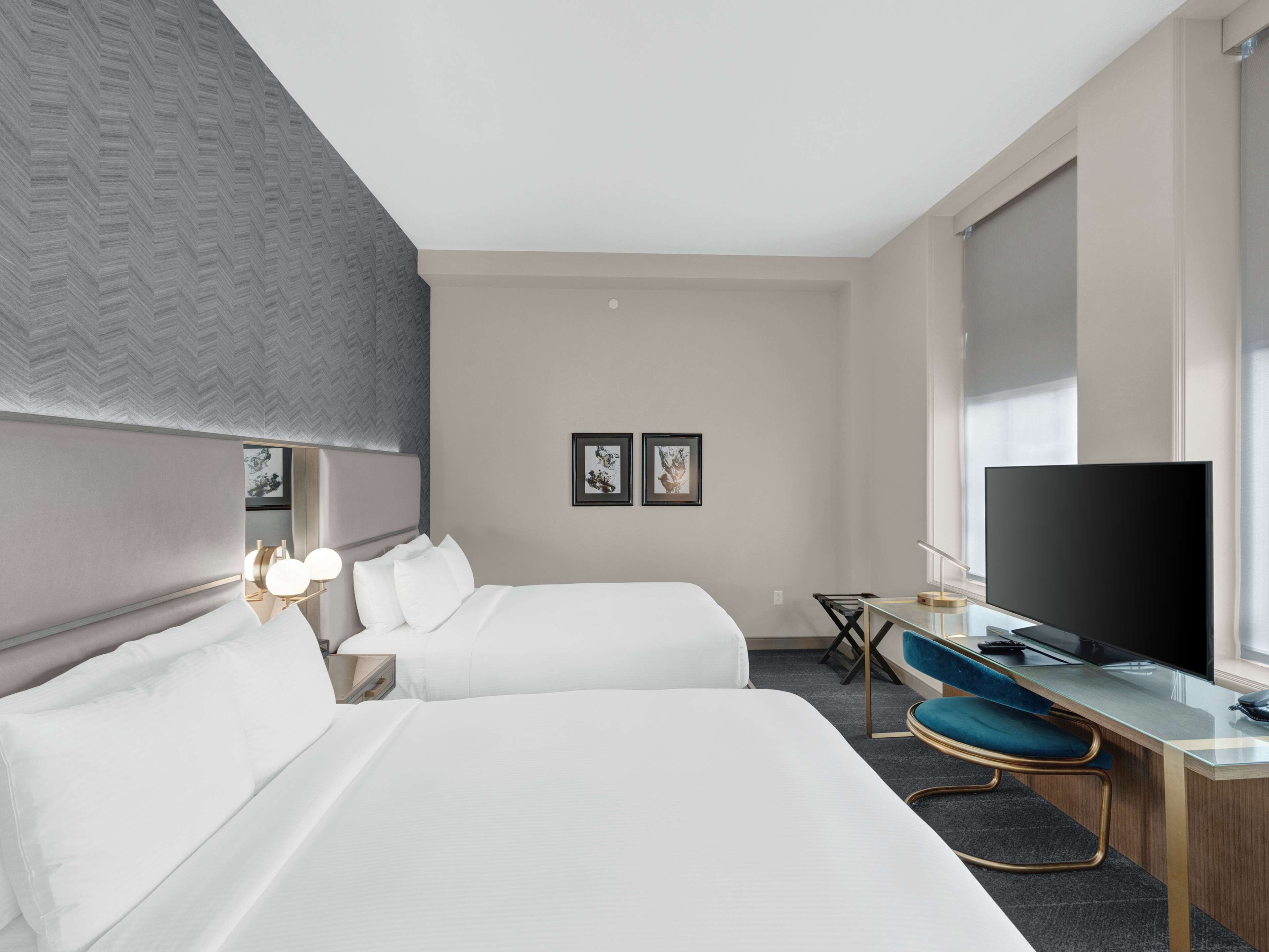 A hotel room with two beds, a desk with a TV, and a large window.