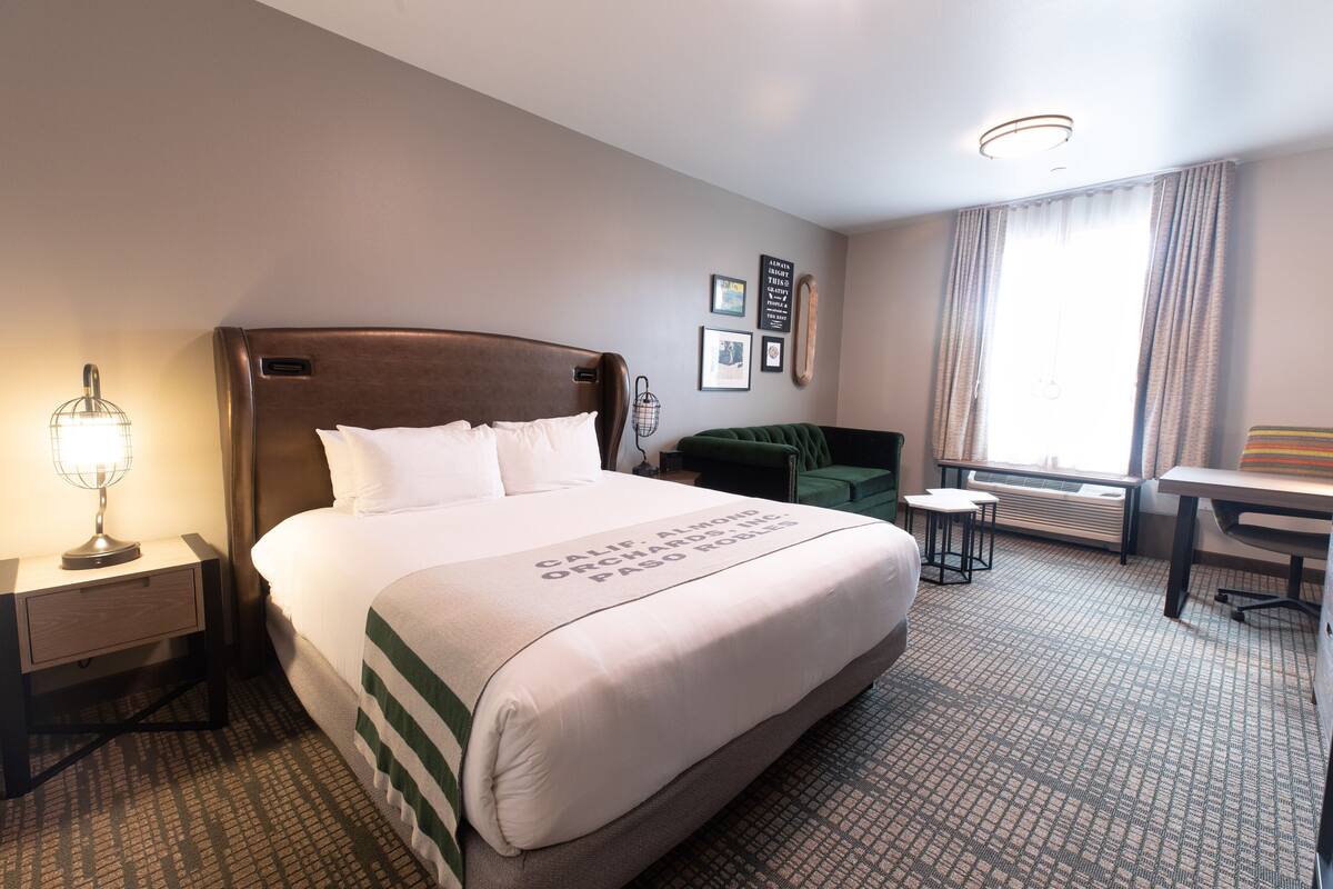 A hotel room with a large bed, a bedside table with a lamp, a small table with two chairs, a green sofa, and a window with curtains.