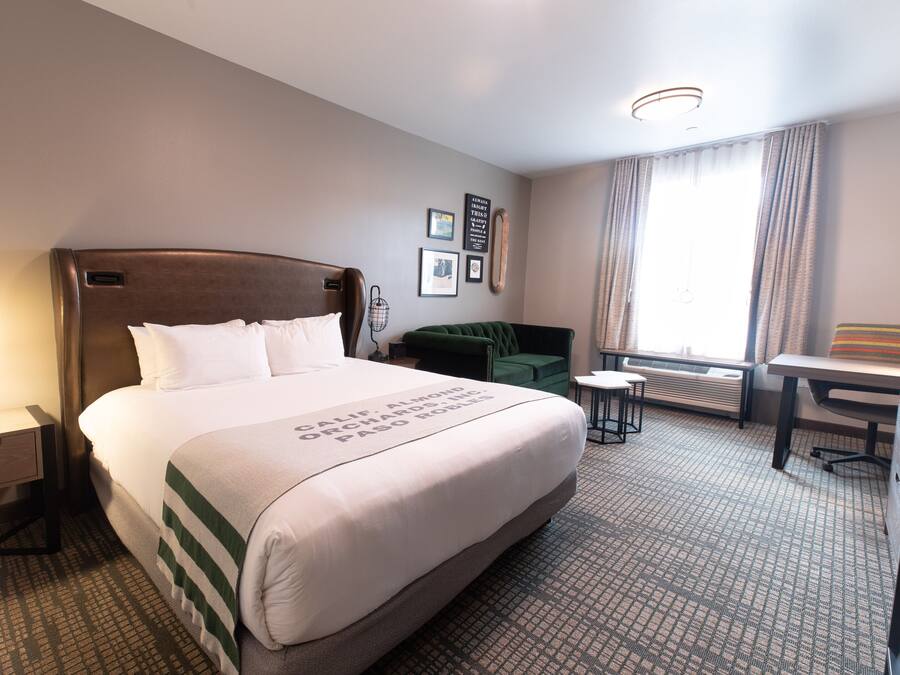 A hotel room with a large bed, a bedside table with a lamp, a small table with two chairs, a green sofa, and a window with curtains.