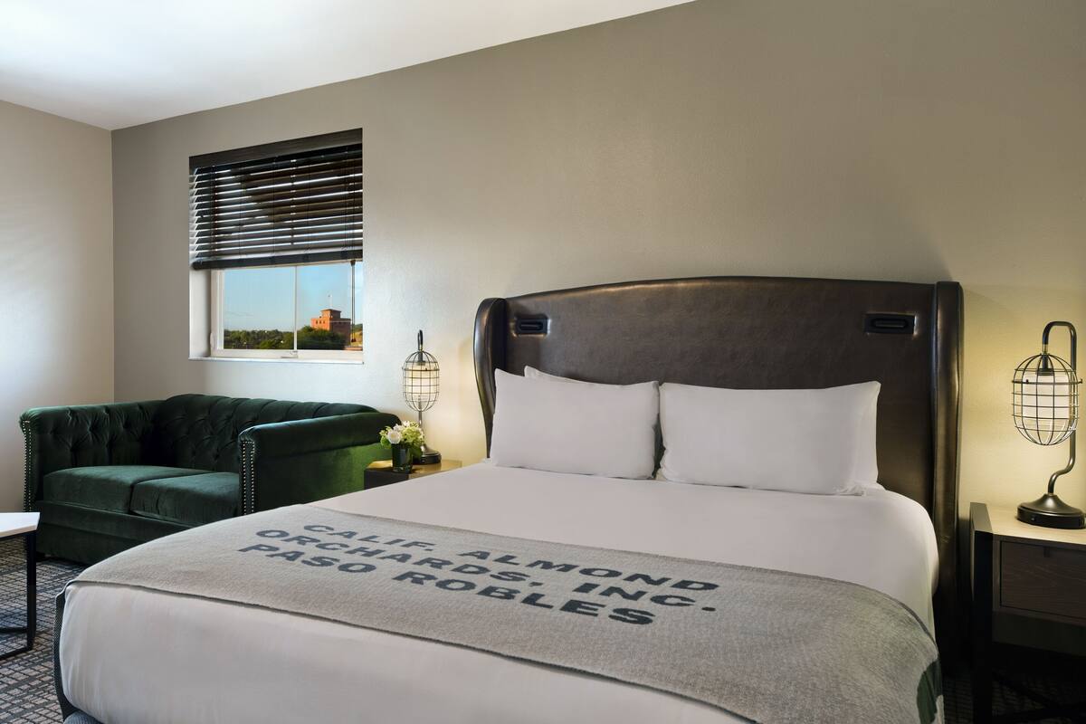 A hotel room with a large bed, a green armchair, a nightstand with a lamp, and a window with blinds.