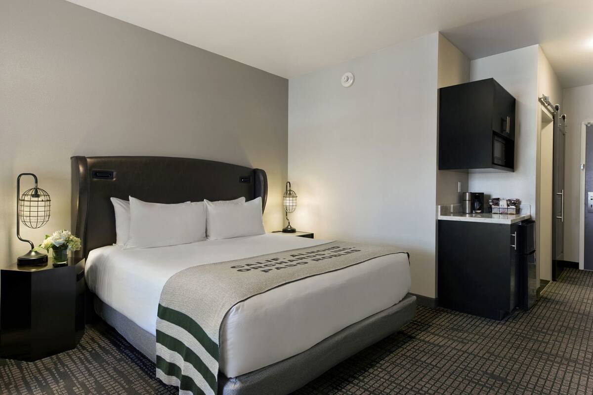 A hotel room with a bed, bedside tables, a nightstand, a desk, a kitchenette, and a door.
