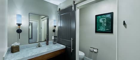 Un baño moderno con lavamanos de mármol, tocador de madera, un espejo grande, un inodoro y un suelo de baldosas hexagonales blanco y negro.