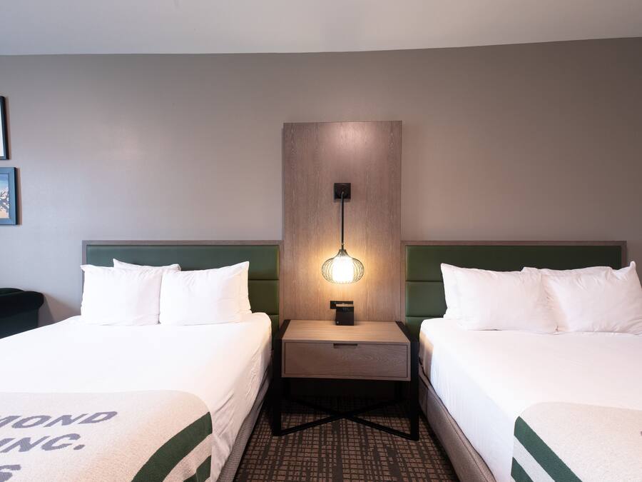 A hotel room with two beds, a nightstand with a lamp, and a wall-mounted light fixture.