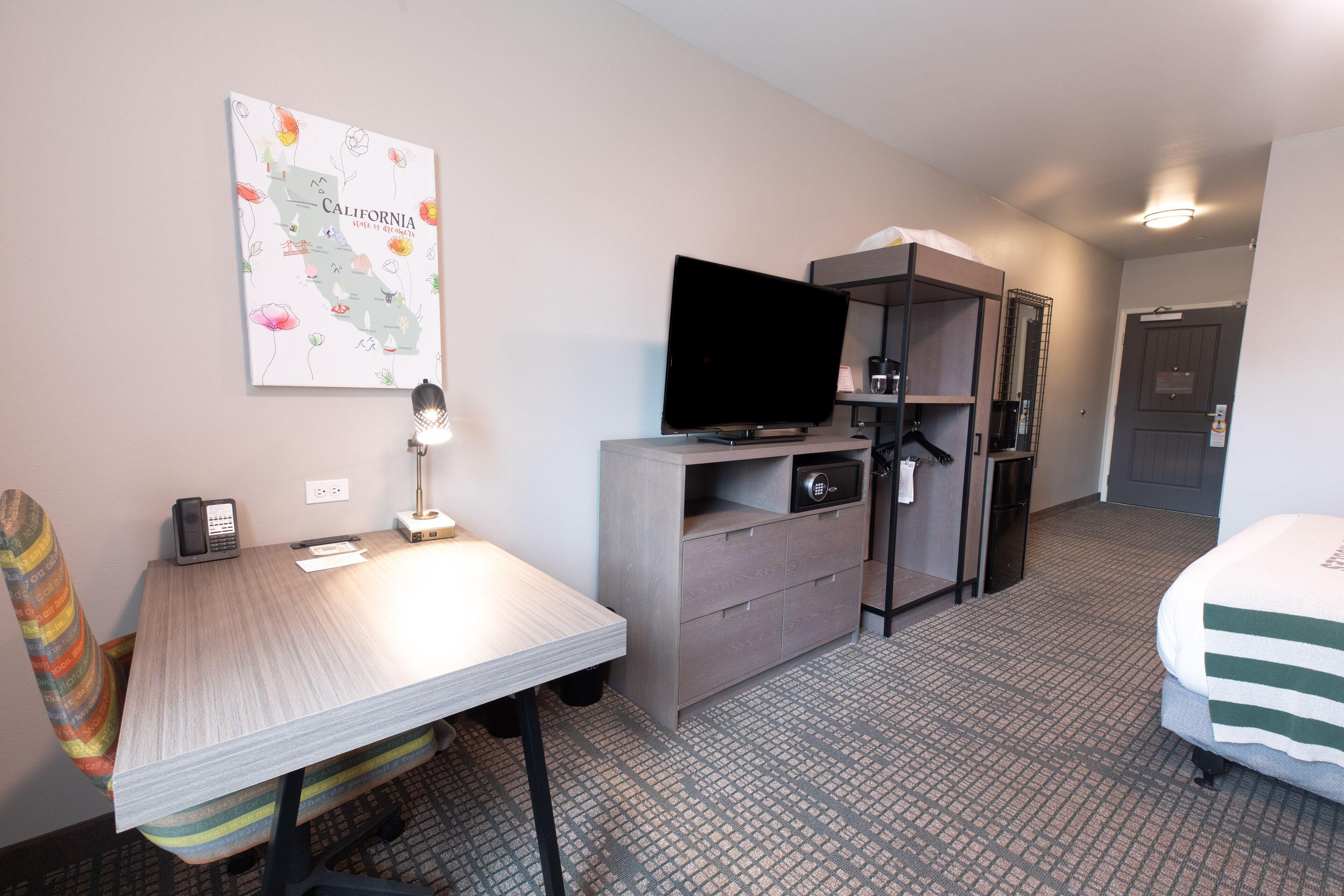 A hotel room with a bed, a desk with a computer, a chair, a television, a wardrobe, and a map on the wall.
