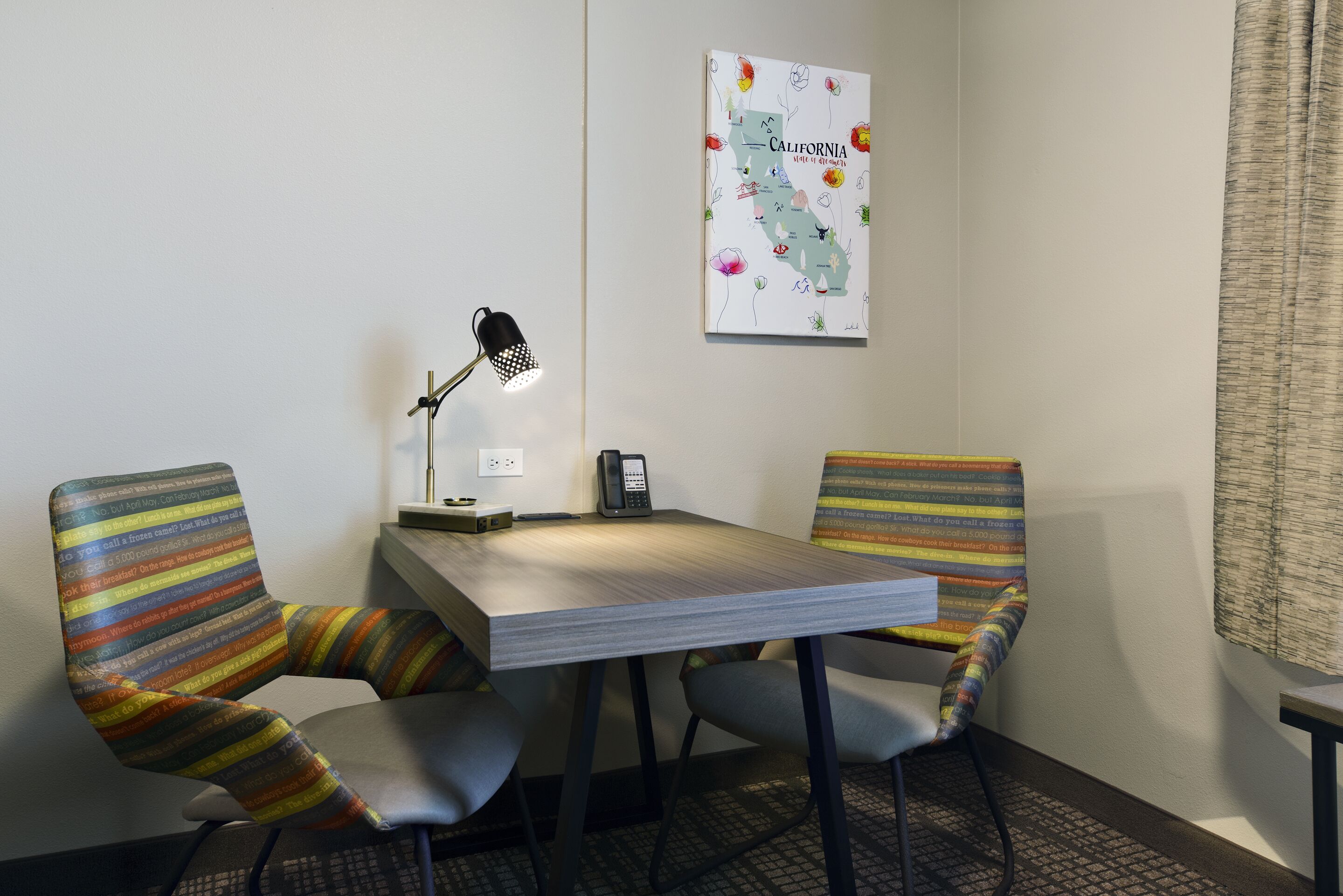 A small, neatly arranged room with a desk, two striped chairs, a lamp, a phone, and a map of California on the wall.