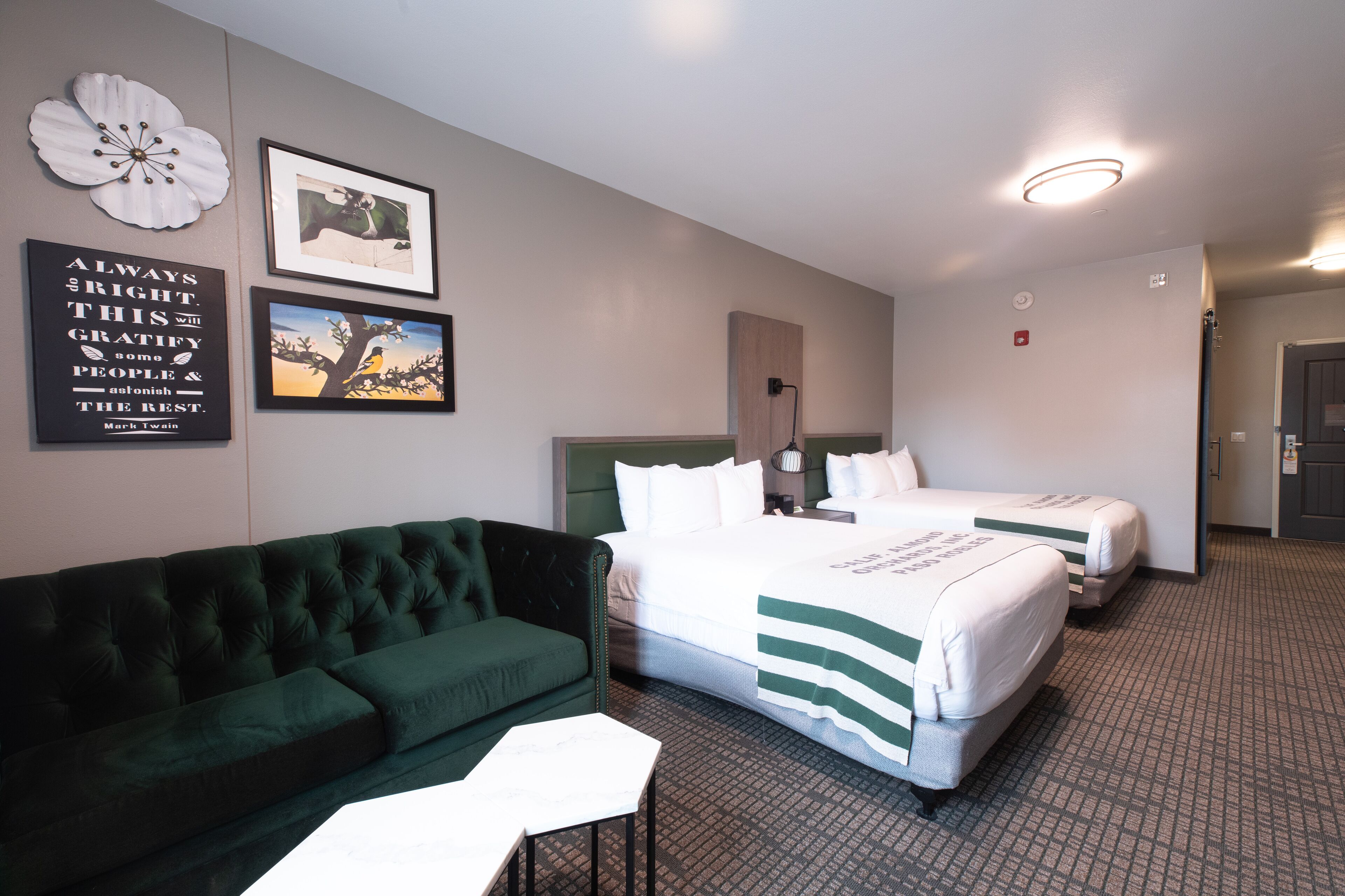 Habitación de hotel con sofá verde, dos camas con ropa blanca, una gran decoración floral y una cita enmarcada en la pared.