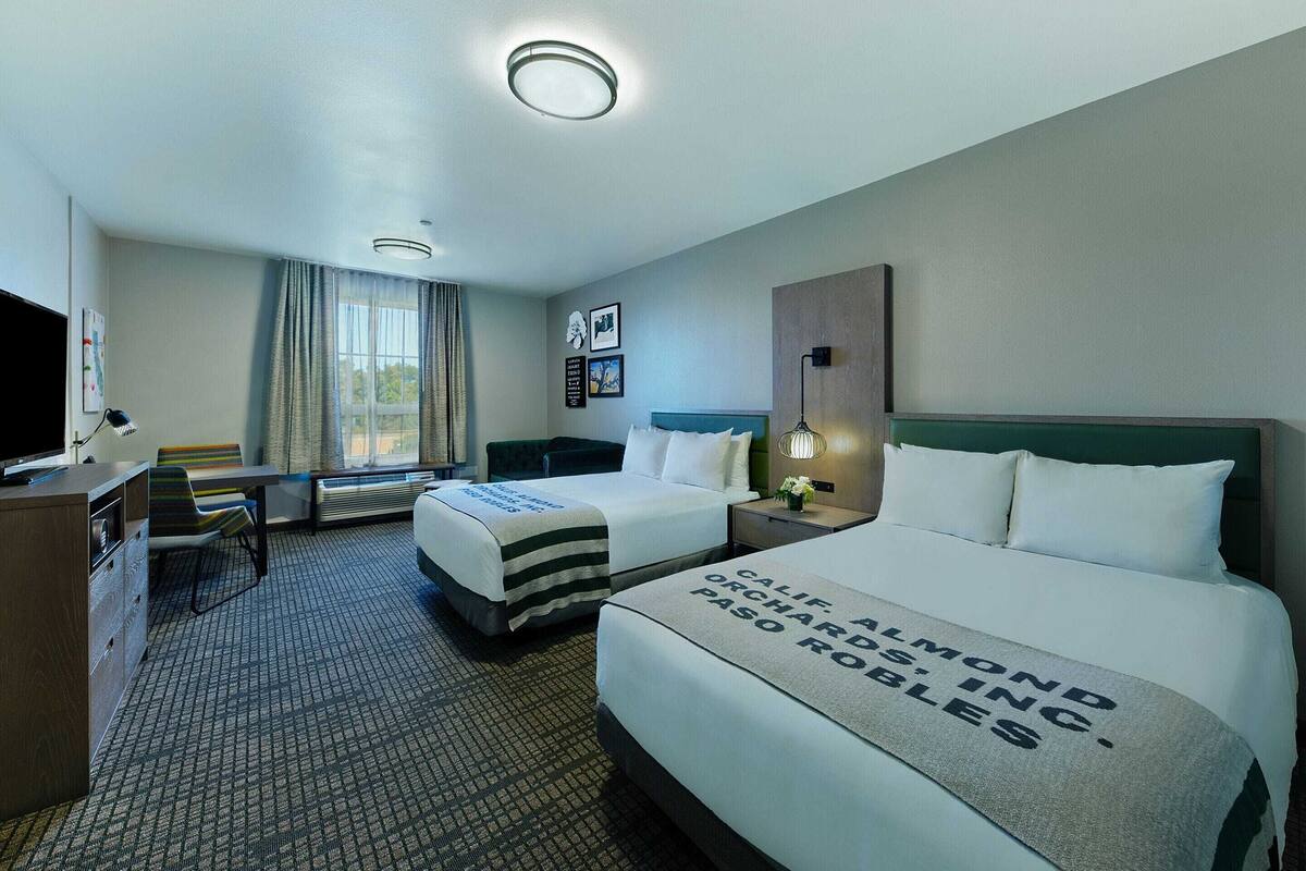 A hotel room with two beds, a desk, a chair, a TV, and a window with curtains.