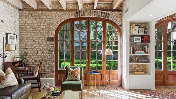 Ein gemütliches Zimmer mit Backsteinwand, Holzbalkendecke, einem grünen Sessel, einem hölzernen Bücherregal und großen Bogenfenstern.