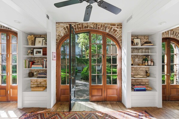 Une pièce dotée d’une grande porte en arc de cercle, d’étagères en bois remplies de livres et d’objets décoratifs, d’un ventilateur de plafond et offrant une vue sur un jardin par la porte.