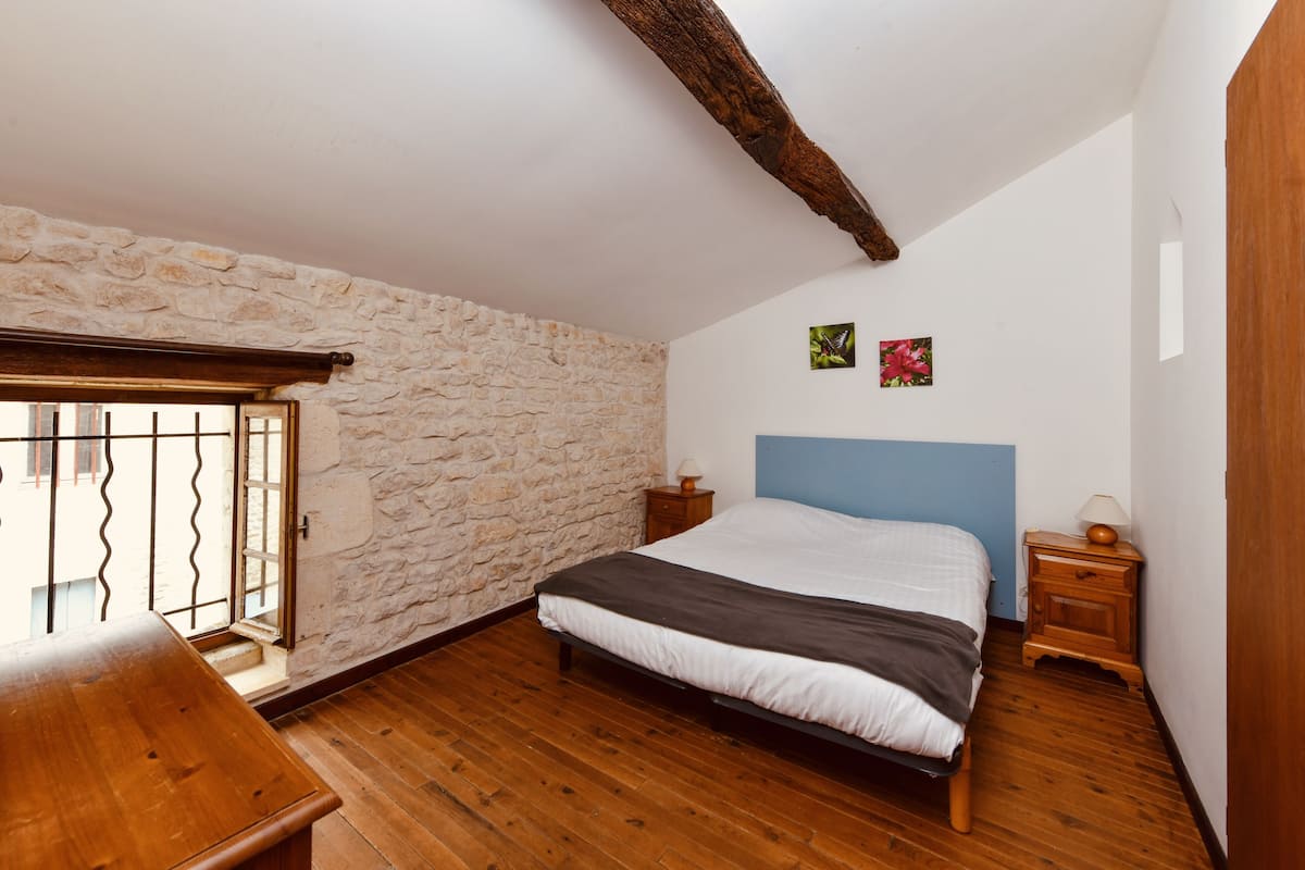 A bedroom with a stone wall, wooden floor, a bed with a blue headboard, a wooden nightstand, and a small table.