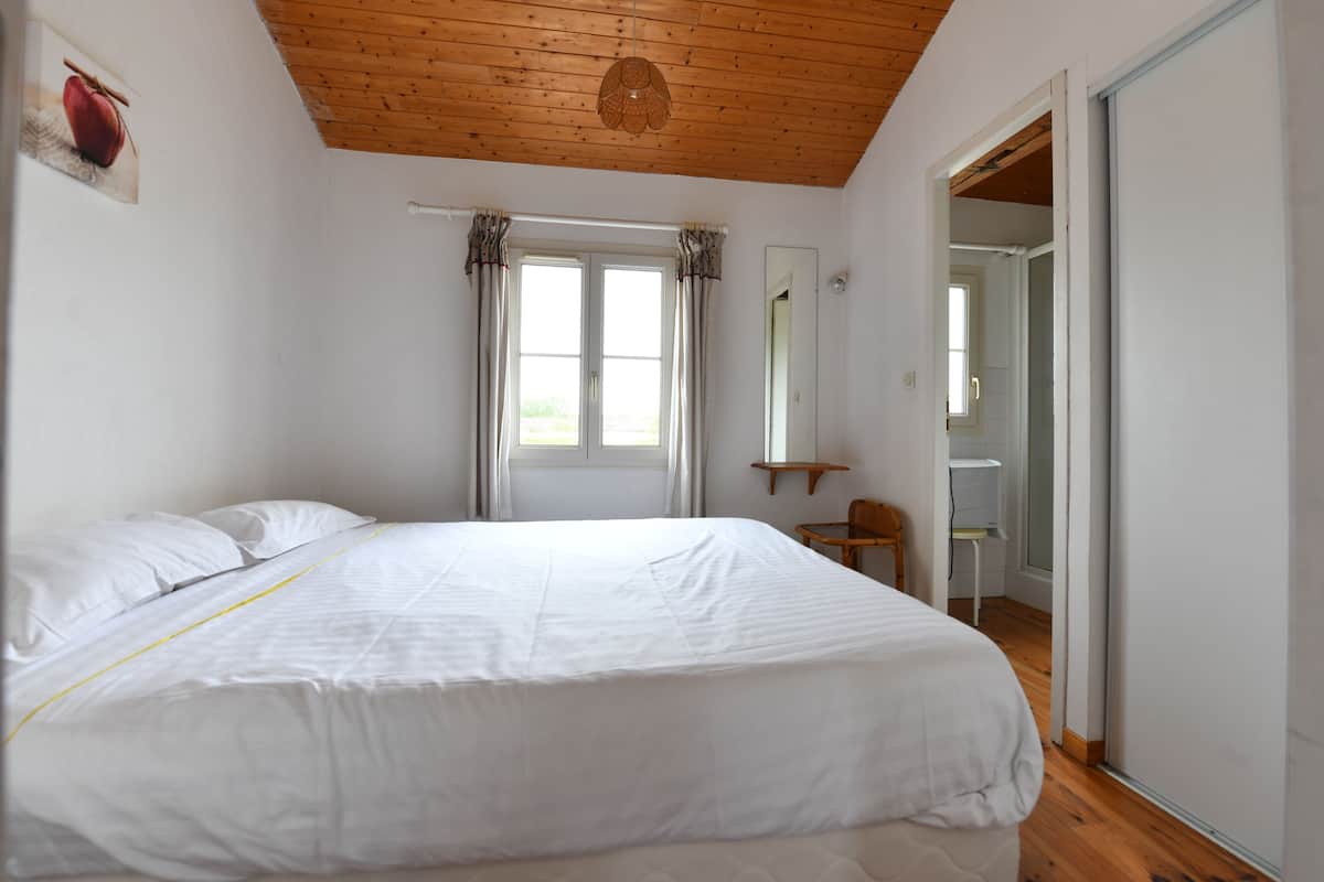 A bedroom with a wooden ceiling, a bed with white bedding, a window with curtains, and a door leading to another room.