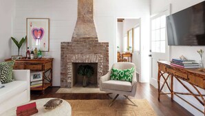 A living room with a brick fireplace, a white sofa, a green and white patterned armchair, a wooden coffee table, and a flat-screen TV mounted on the wall. - Green Cottage – Private Casa Zoë Downtown Stay (Charleston)