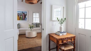 A hallway with a white door, a wicker lamp, a white sofa with cushions, a round coffee table, and a small wooden table with a vase and books. - Green Cottage – Private Casa Zoë Downtown Stay (Charleston)