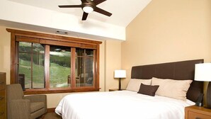 A bedroom with a large bed, a ceiling fan, a window with a view of a grassy area, a chair, and a bedside table with a lamp. - Alpine Condo + Fireplace, Balcony | Access to Spa (Breckenridge)