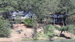 A lakeside cabin with a deck and multiple windows, surrounded by dense trees. - Top Lakefront Oasis - Kayaks, Paddle Bd, - Shasta Lakeshore Retreat #2 (Lakehead)