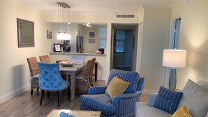 A living room with a sofa, armchair, and coffee table. There is a dining area with chairs and a table. A kitchen is visible in the background. - Stunning Beachfront Condo in Gated Community (Marco Island)