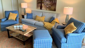 A living room with striped blue furniture, a glass coffee table, and two lamps. - Stunning Beachfront Condo in Gated Community (Marco Island)
