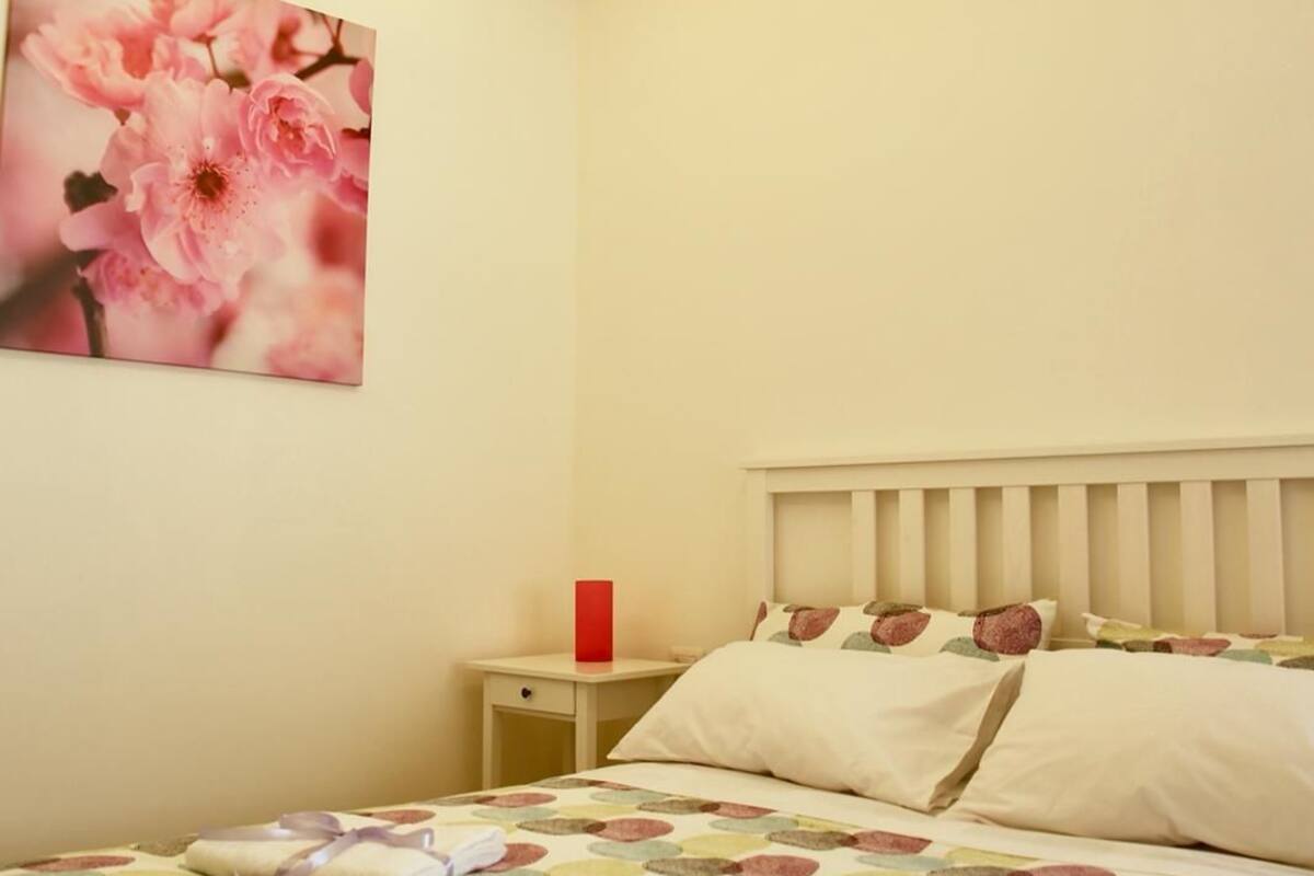 Un lit bien fait, recouvert d’une couvre-lit aux motifs circulaires et colorés, avec un oreiller aux motifs rouges et bruns, et une table de chevet sur laquelle se trouve un objet rouge.