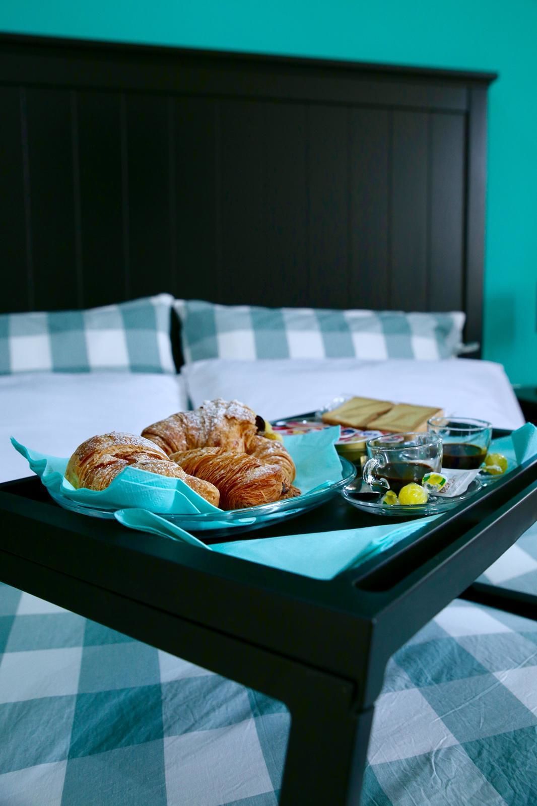 Ein Bett mit kariertem Überwurf, ein Tablett mit Croissants, ein Glas Wasser und eine Tasse Kaffee auf einem Nachttisch.