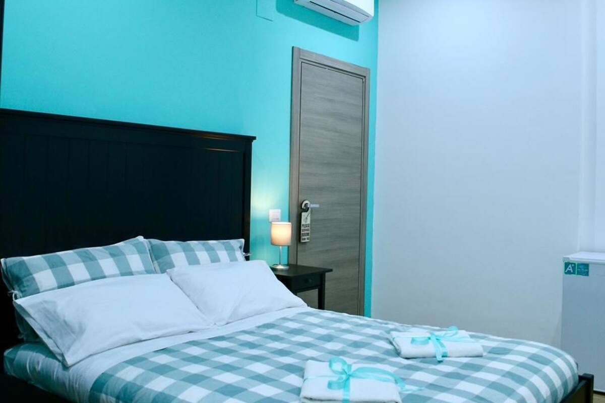 Une chambre moderne avec une couvre-lit à carreaux, une tête de lit sombre, un climatiseur, une table de chevet avec une lampe et une porte dotée d’une poignée.