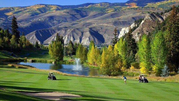 Un campo de golf con un estanque, árboles y montañas al fondo.