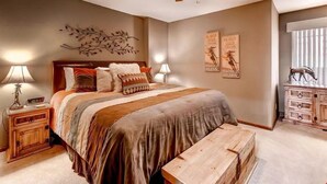 Un dormitorio con cama, mesitas de noche, un banco de madera, ventilador de techo, decoración en la pared y una ventana con persianas.