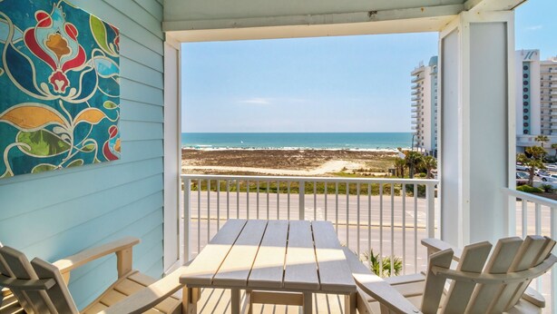Un balcon avec vue sur la plage et l’océan, des meubles en bois et une peinture abstraite aux couleurs vives accrochée au mur.