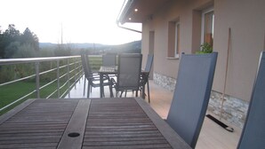 Eine Holzterrasse mit Tisch und Stühlen, einem Metallgeländer und Blick auf eine grüne Landschaft.