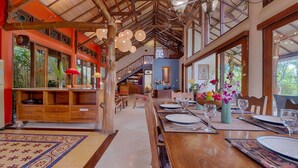 A spacious dining area with a long wooden table set for a meal, surrounded by large windows offering a view of the outdoors. - Vibrant Soulful Bali Home for Short Term Rent (North Sanur)