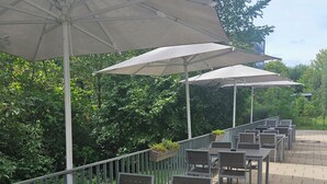 Un espace extérieur aménagé en terrasse, avec plusieurs tables et chaises, dont certaines sont placées sous des parasols, le tout entouré de verdure.
