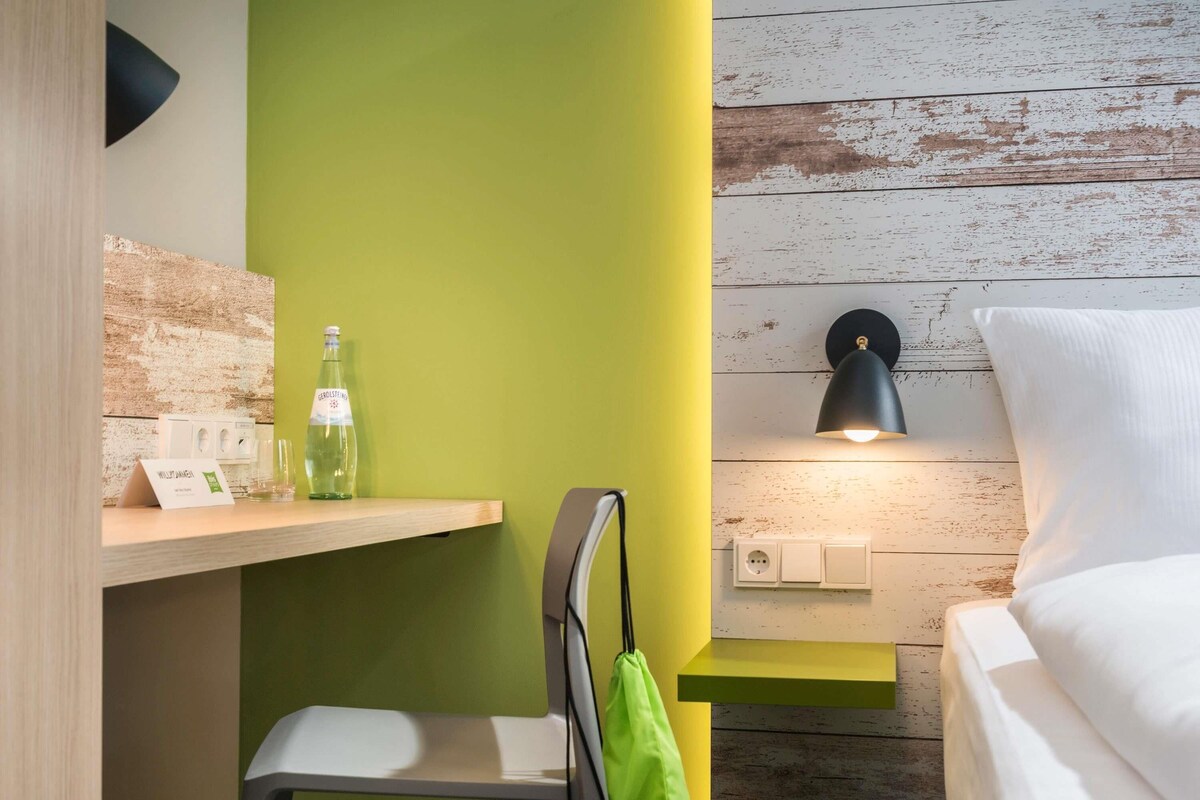 Habitación de hotel con escritorio de madera, pared de acento verde, cama con ropa blanca, lámpara de escritorio negra y toalla verde colgada de la silla.