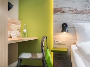 Habitación de hotel con escritorio de madera, pared de acento verde, cama con ropa blanca, lámpara de escritorio negra y toalla verde colgada de la silla.