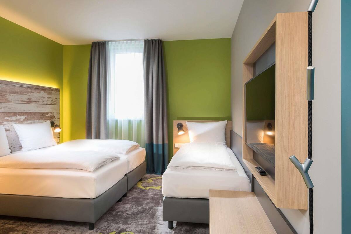 A hotel room with two beds, a desk, a TV, and a window with curtains.