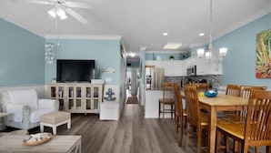 A spacious living room with a television, a dining area with wooden chairs, and a kitchen with white cabinets. - Cute & Beachy! Updated condo! 2 bed/2 bth condo/200 steps to beach! (Tybee Island)