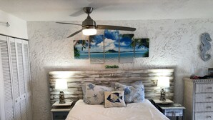 Un dormitorio con cabecera de madera, ventilador de techo, cama con colcha de rayas azules y blancas, y pared decorada con un mural de playa tropical.