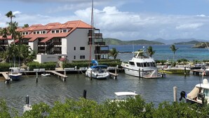 Un puerto deportivo con varias embarcaciones amarradas, un edificio de techo rojo y vistas al mar y colinas al fondo.