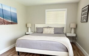 Une chambre à coucher comprenant un lit, deux tables de chevet, une fenêtre avec des stores, une œuvre d’art accrochée au mur et un panneau décoratif encadré.