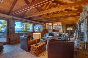 A rustic cabin interior with a wooden ceiling, a stone fireplace, and a large window overlooking a forested area.