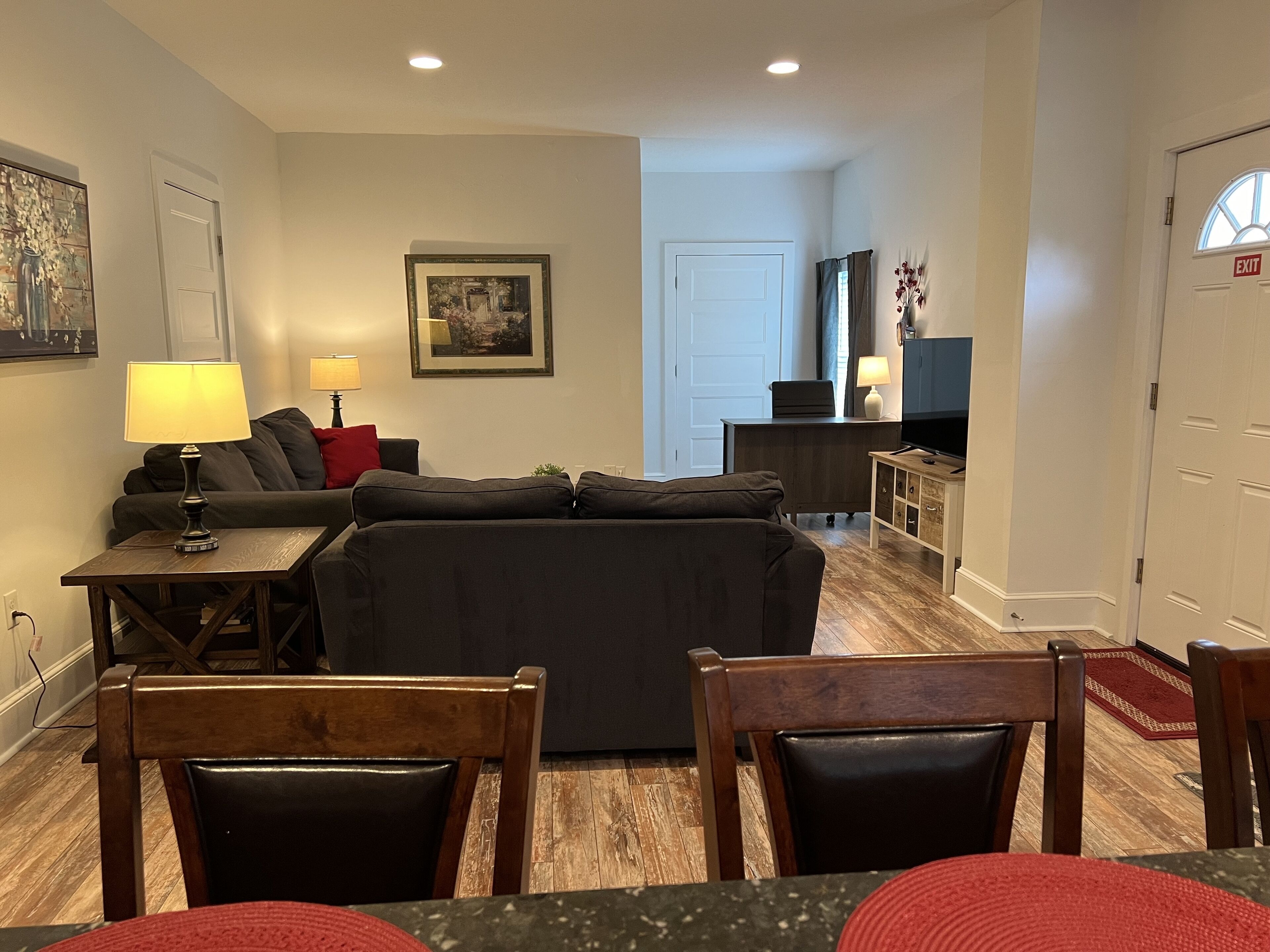 A living room with a sofa, a coffee table, and a TV. There is a door with an exit sign, a lamp, and a framed picture on the wall.