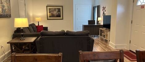 A living room with a sofa, a coffee table, and a TV. There is a door with an exit sign, a lamp, and a framed picture on the wall.