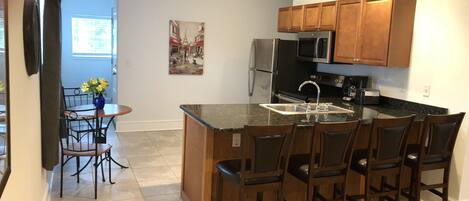 A modern kitchen with wooden cabinets, a stainless steel refrigerator, a microwave, and a sink. There is a dining area with a table and chairs.