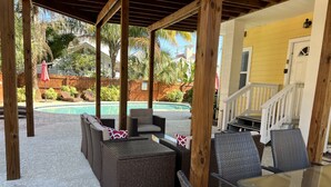 Un patio cubierto con mobiliario de mimbre y una mesa de vidrio, con vistas a una piscina y un edificio amarillo.