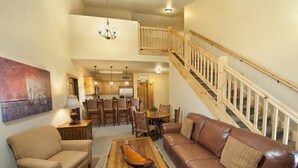 A spacious living room with a brown leather sofa, a wooden coffee table, and a dining area with wooden furniture. A staircase with a wooden railing leads to an upper level.
