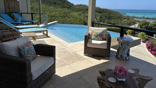 Ein Poolbereich mit Liegestühlen, einer Korbsesselcouch und Blick auf das Meer und die Hügel.