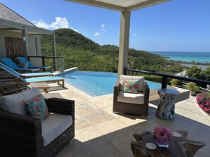 Un espace aménagé au bord de la piscine, avec des chaises longues, un canapé en osier et une vue sur l’océan et les collines.